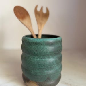 May include: A cylindrical ceramic utensil holder with a textured, wavy surface finished in a deep forest green glaze. The base reveals a natural, unglazed tan clay. Two wooden utensils, a spoon and a fork, are placed inside the container.