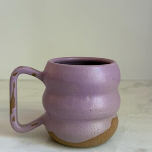 May include: A handmade ceramic mug with a unique, textured design. The mug features a lavender-colored body with a natural, tan-colored base. The handle has a matching lavender hue with tan accents. This mug is perfect for coffee, tea, or any beverage.