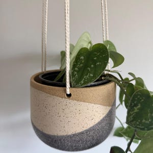 May include: A hanging ceramic planter with a three-tone design featuring tan, cream, and dark gray sections. The cream section has a speckled texture, while the dark gray base has a rougher finish. A trailing heart-shaped plant grows inside.