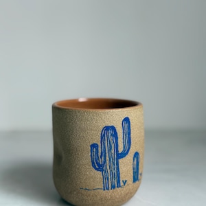 May include: A tan ceramic cup with a blue cactus illustration. The cup has a textured surface and a brown interior. The cactus design features a large central cactus and a smaller one to the right.