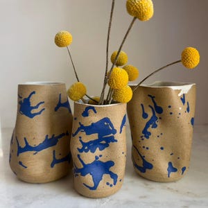 May include: Three ceramic vases with a speckled tan finish and irregular, hand-formed shapes. Each vessel features a vibrant blue paint splatter pattern across the exterior. The center vase holds several dried yellow billy ball flowers.