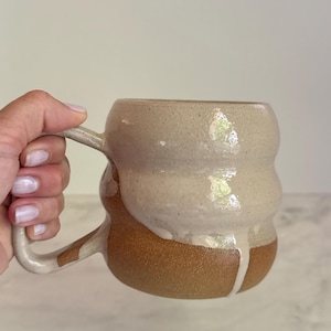 May include: Handmade ceramic mug with a unique, rounded shape. The upper portion is a creamy white glaze, flowing down to meet a warm, earthy brown base. The mug has a sturdy handle for easy gripping.