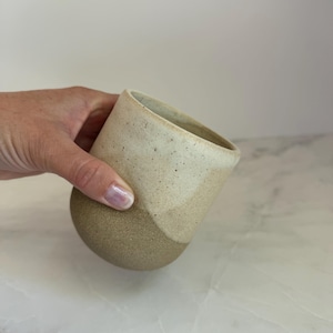 May include: A ceramic tumbler featuring a two-tone design with a speckled cream-colored upper section and a solid, textured brown base. The cup has a rounded, handleless silhouette with a smooth rim and a matte finish on the lower portion.