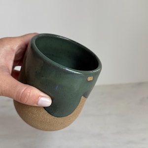 May include: A handmade ceramic mug with a dark green glazed top and a tan, unglazed bottom. The mug has a unique, asymmetrical shape and a small gold accent. The mug is held by a person's hand.