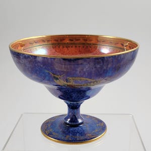 May include: A blue and gold footed bowl with a wide rim. The bowl has a textured, iridescent finish. The interior is a reddish-orange color with gold detailing. The base and rim are trimmed with gold accents.