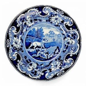 Könnte beinhalten: Ein blauer und weißer Porzellanteller mit einem floralen Rand und einem zentralen Bild eines Hundes, der durch ein Waldgebiet rennt.
