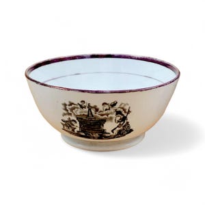 May include: Cream-colored ceramic bowl with a decorative black and white illustration. The bowl has a wide rim with a dark purple border and a pedestal base. The illustration depicts figures and architectural elements.