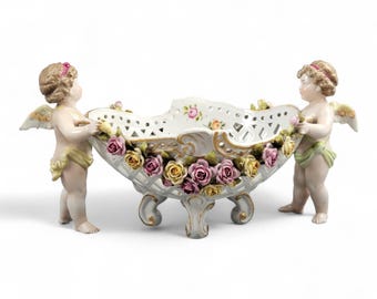 May include: A white porcelain centerpiece bowl with a lattice design, supported by three ornate legs. Two cherubic figures with wings flank the bowl, which is adorned with pink and yellow roses. Gold accents highlight the details.