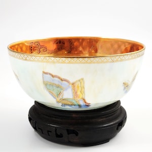 May include: A decorative porcelain bowl with a gold rim and butterfly motif. The bowl is supported by a dark wooden stand. The interior of the bowl has a gold design. The butterflies are painted in shades of blue, yellow, and green.