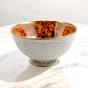 May include: A white ceramic bowl with a gold rim and a geometric pattern. The inside of the bowl is a reddish-brown color.
