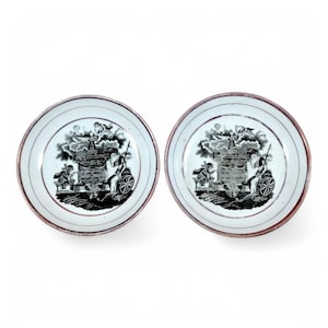 May include: Two white porcelain saucers with black and red trim. Each saucer features a black and white image of a cart pulled by a horse with a figure standing on top of the cart.