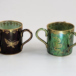 May include: Two decorative ceramic mugs with gold trim and handles. One is dark with a gold butterfly, the other is green with a gold snake design. Both have gold accents on the rim.