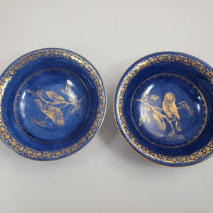 May include: Two matching blue ceramic bowls with gold trim and bird and branch designs. The bowls have a dark blue interior and a gold floral pattern around the rim. The bird and branch design is in gold.