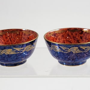 May include: Two ceramic bowls. The bowls have a blue exterior and a red interior. They feature a gold rim and a gold dragon design. The bowls are displayed on a clear surface.