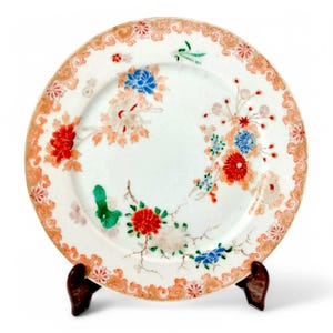 May include: A white porcelain plate with a gold rim and floral designs in red, orange, blue, and green. The plate is on a wooden stand.