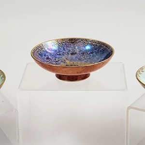May include: Three decorative ceramic bowls with iridescent interiors and gold rims. The bowls have a dark blue base and a light blue, copper, and blue interior. The bowls are displayed on clear acrylic stands.