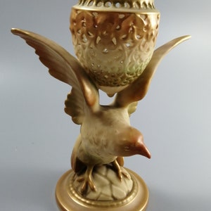May include: A ceramic bird figurine with outstretched wings supporting a decorative vase. The vase has a pierced design and a gold rim. The bird and base are in shades of brown and gold, creating an elegant, vintage aesthetic.