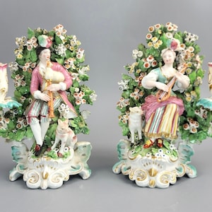 Op de afbeelding: Set van porseleinen beeldjes met een man die doedelzak speelt en een vrouw met een luit, elk met een hond, tegen een bloemenachtergrond. Elke figuur heeft een kandelaar. De kleuren zijn roze, groen, wit en goud.