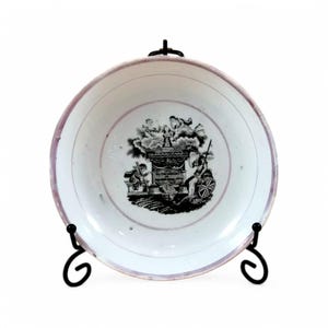 May include: A white ceramic bowl with a black and white design of a building with a chimney and two figures. The bowl has a pink rim and is on a black stand.
