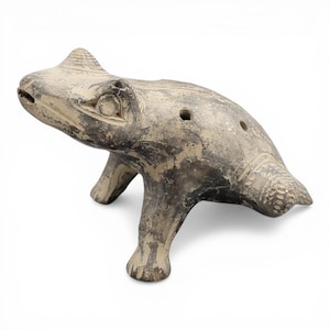 Puede incluir: Ocarina de cerámica en forma de rana, en tonos grises y beige. El instrumento tiene una superficie texturizada, tres agujeros para los dedos y una cara detallada de rana. La ocarina está sobre cuatro patas.