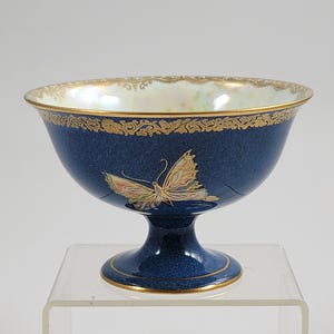 May include: A blue footed bowl with a butterfly design. The bowl has a gold rim and a gold decorative pattern around the top. The butterfly is in the center of the bowl and has pink, yellow, and gold accents.