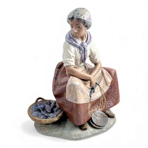 Peut inclure: Figurine en céramique d'une femme assise, vêtue d'une blouse blanche, d'une écharpe violette et d'une longue jupe brun-rouge. Elle tient une balance et est assise à côté d'un panier de poissons. La figurine est sur une base en forme de pierre.