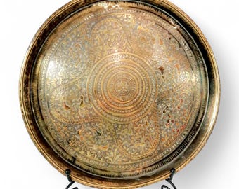 May include: A round, gold-colored metal tray with intricate engraved designs. The tray is on a black metal stand with three legs.