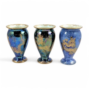 May include: Three iridescent blue vases with gold dragon motifs. Each vase has a flared rim and a pedestal base, with gold detailing. The vases are arranged in a row against a white backdrop.