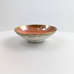 May include: A small, white ceramic bowl with a gold rim and a red and gold speckled interior. The bowl has a delicate, scalloped edge.