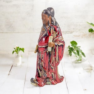 May include: A ceramic figurine of a man in a red and black robe holding a scroll. The figurine is standing on a white wooden surface.