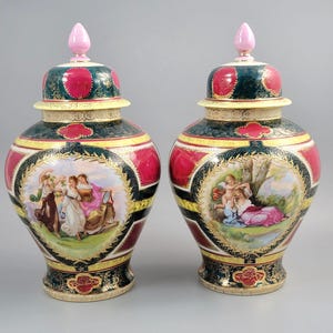 Op de afbeelding: Twee decoratieve keramische vazen met deksels. De vazen hebben een rood, groen en goud kleurenschema met geschilderde scènes van figuren. Elke vaas heeft een roze finial op het deksel en een gedetailleerde basis.