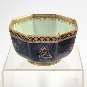 May include: An antique, hexagonal bowl with a dark blue exterior and a light green interior. The bowl features gold trim along the rim and base, with gold floral designs. The bowl is displayed on a clear acrylic stand.