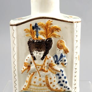 Puede incluir: Una botella de cerámica blanca con cuerpo rectangular y cuello cilíndrico pequeño. El frente presenta un diseño en relieve de dos figuras con vestimenta de época, con detalles en marrón, dorado y azul. La botella tiene un borde decorativo.