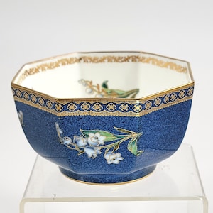 May include: An octagonal blue ceramic bowl with a textured surface. The bowl has a gold rim and floral designs with white flowers and green leaves. The interior is white with gold detailing.