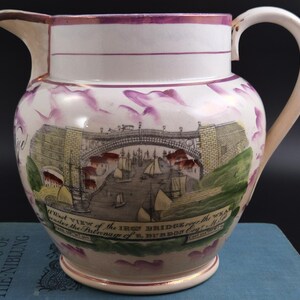 May include: A ceramic jug featuring a transfer print of the Iron Bridge over the River Wear. The design includes a detailed bridge, sailing ships, and text reading A West View of the IRON BRIDGE over the WEAR. It has purple lustre accents.