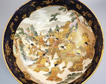 Könnte beinhalten: Dekorative Keramikschale mit einer detaillierten japanischen Szene. Die Schale hat einen dunkelblauen Rand mit goldenen Akzenten und ein zentrales Bild von Kriegern in der Schlacht. Das Kunstwerk zeigt Figuren in Rüstungen, Bäume und eine Landschaft in leuchtenden Farben.