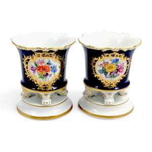 Könnte beinhalten: Paar Porzellan-Blumentopf mit Kobaltblau-Glasur und goldenen Akzenten. Jeder Topf zeigt ein Blumenmuster mit einem herzförmigen Rahmen. Die Töpfe haben drei Beine und einen gewellten Rand.