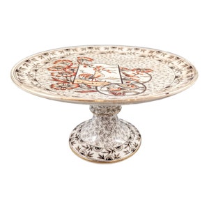 May include: A white ceramic cake stand with a gold rim and a floral design. The stand features a central design of a bird and a fish.