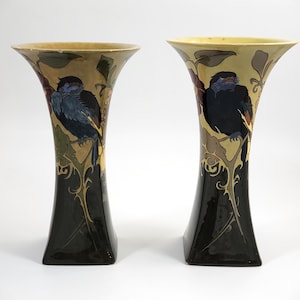 May include: Two decorative vases with a flared top and a black base. Each vase features a bird perched on a branch with leaves and floral accents. The color palette includes black, gold, blue, and brown.