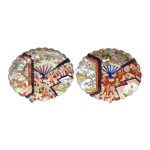 May include: Two decorative, scalloped-edge plates with intricate Japanese-style scenes. Each plate features a red central section with figures, a blue fan design, and detailed landscape and floral elements in various colors.