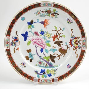 May include: A white porcelain plate with a floral design. The plate features a pink peony, blue and yellow flowers, and brown branches. The rim has a brown and red pattern with a gold trim. Butterflies and other decorative elements are also present.