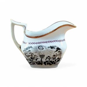 May include: A white ceramic creamer with a brown and black design of angels playing music around a central figure. The creamer has a pink and brown trim around the top.