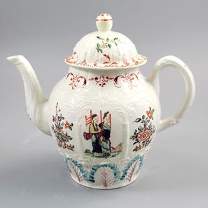 May include: A white porcelain teapot with a lid, spout, and handle. The teapot features floral and foliate designs in red, green, and blue. Two figures are depicted in a central panel. The teapot has a bulbous body and a decorative lid.