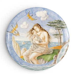 May include: Round decorative plate featuring a serene scene of a woman cradling a child, set against a coastal backdrop. The plate has a blue border with Greek lettering and includes images of birds, clouds, and a waterfall. The artwork is rendered in soft, muted colors.