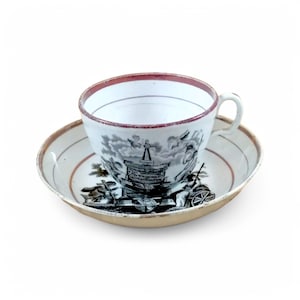 May include: A vintage teacup and saucer set with a black and white illustration of a ship and a red rim. The saucer has a gold rim.