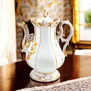 May include: A white porcelain teapot with gold accents and a delicate floral design. The teapot has a rounded body, a spout, a handle, and a lid. The teapot is sitting on a wooden table.
