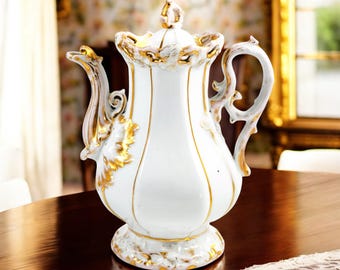 May include: A white porcelain teapot with gold accents and a delicate floral design. The teapot has a rounded body, a spout, a handle, and a lid. The teapot is sitting on a wooden table.