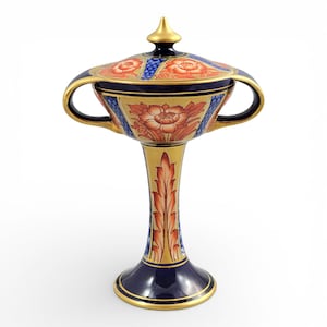 May include: An ornate ceramic urn with a lid, decorated with gold, navy blue, and orange floral patterns. The urn has two handles and a tall, slender base. The lid is topped with a gold finial. The design is intricate and detailed.
