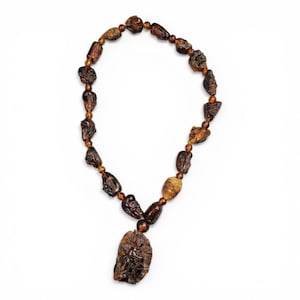 Peut inclure: Un collier de perles ambrées avec un pendentif. Les perles sont de formes et de tailles variées, certaines sculptées. Le pendentif présente une sculpture détaillée de personnages. Le collier est présenté sur fond blanc.