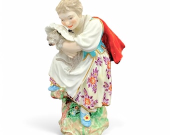 Op de afbeelding: Een porseleinen beeldje van een meisje in een witte jurk met een bloemenpatroon, dat een klein hondje vasthoudt. Ze draagt een blauw en gouden lijfje en een rode cape. Het beeldje staat op een groene basis met bloemen.